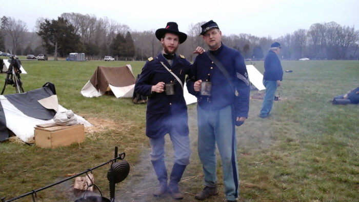 Two men dressed as Civil War soldiers wearing blue uniforms and holding tin cups stand in a field with small tents and a camp fire.