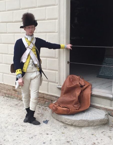 A man dressed as a Revolutionary War soldier in a blue coat with yellow trim and a black hat with a piece of fure on the top stands beside a masonry building with a leather bag near his feet.